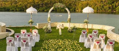 An outdoor wedding setup with a lake view, white chairs with red sashes, and a floral arch.