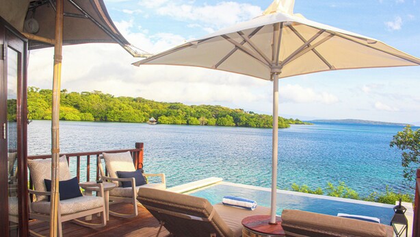 Una terraza de madera con sillones de descanso y un gran paraguas, con vista a un mar azul cristalino y abundante vegetación.