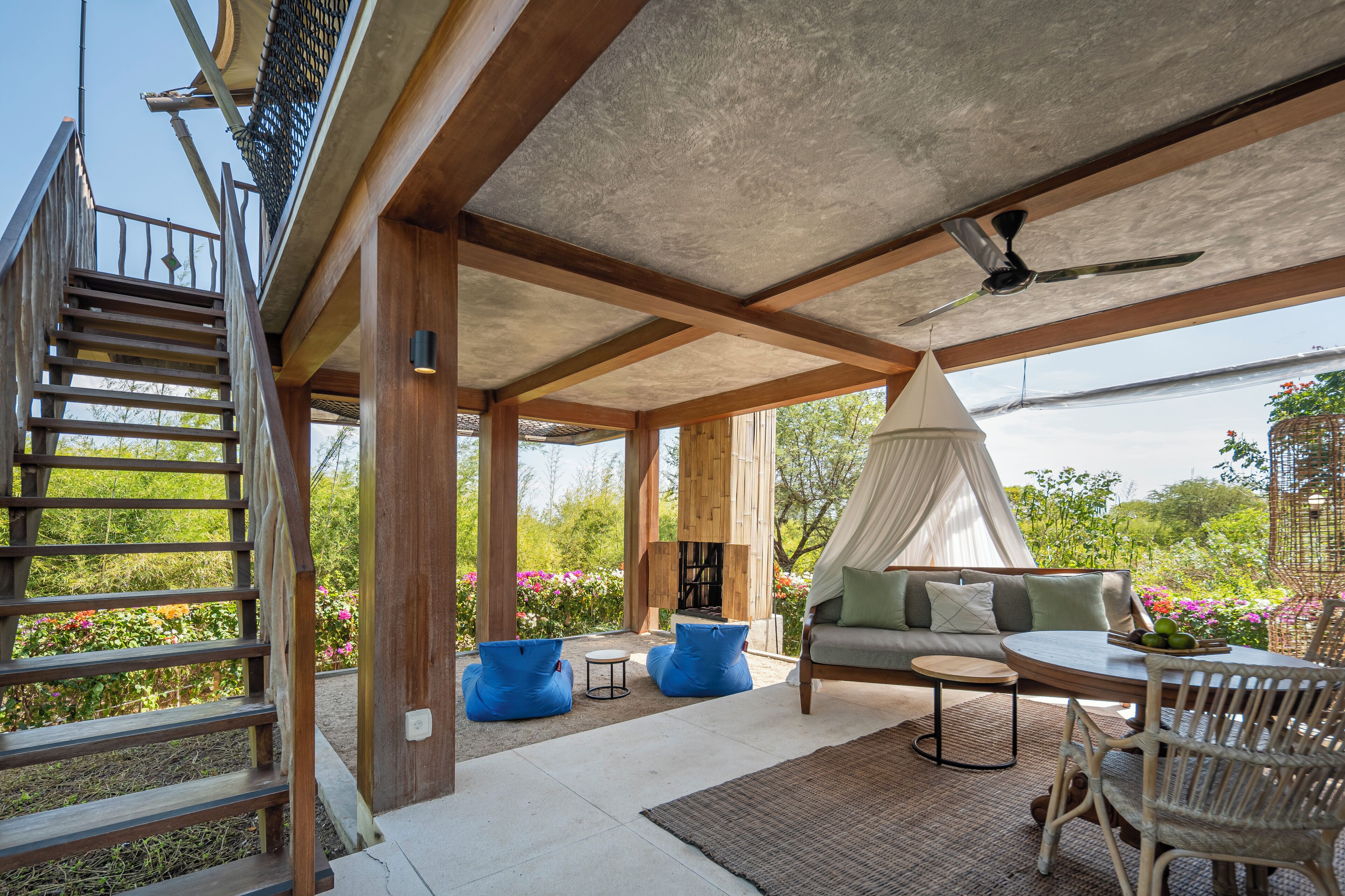 A covered outdoor area with a sofa, chairs, and a table. There is a wooden staircase and a fire pit.