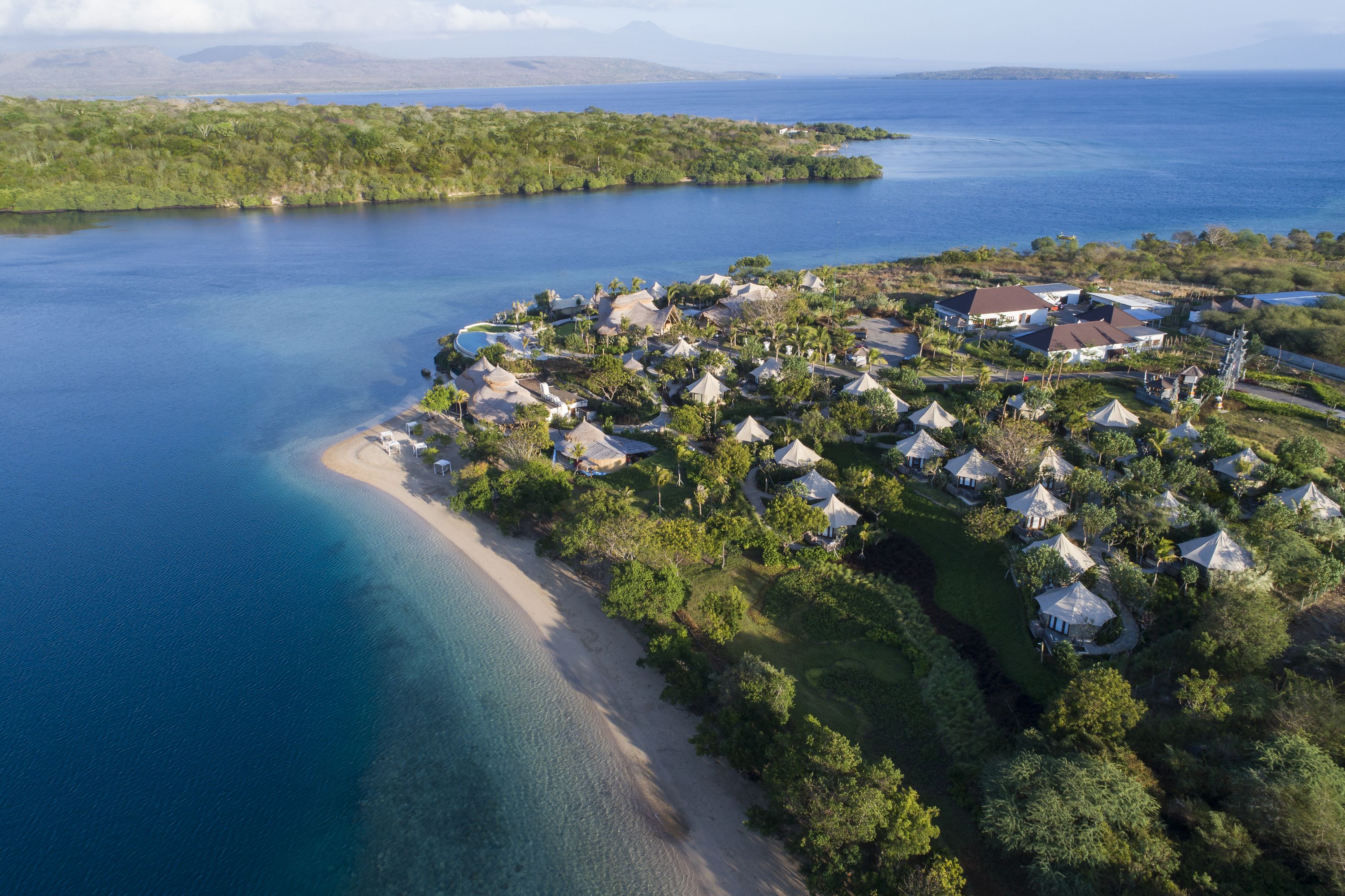 An aerial view of a coastal resort with multiple bungalow-style huts, a sandy beach, and a backdrop of lush greenery and a large body of water.