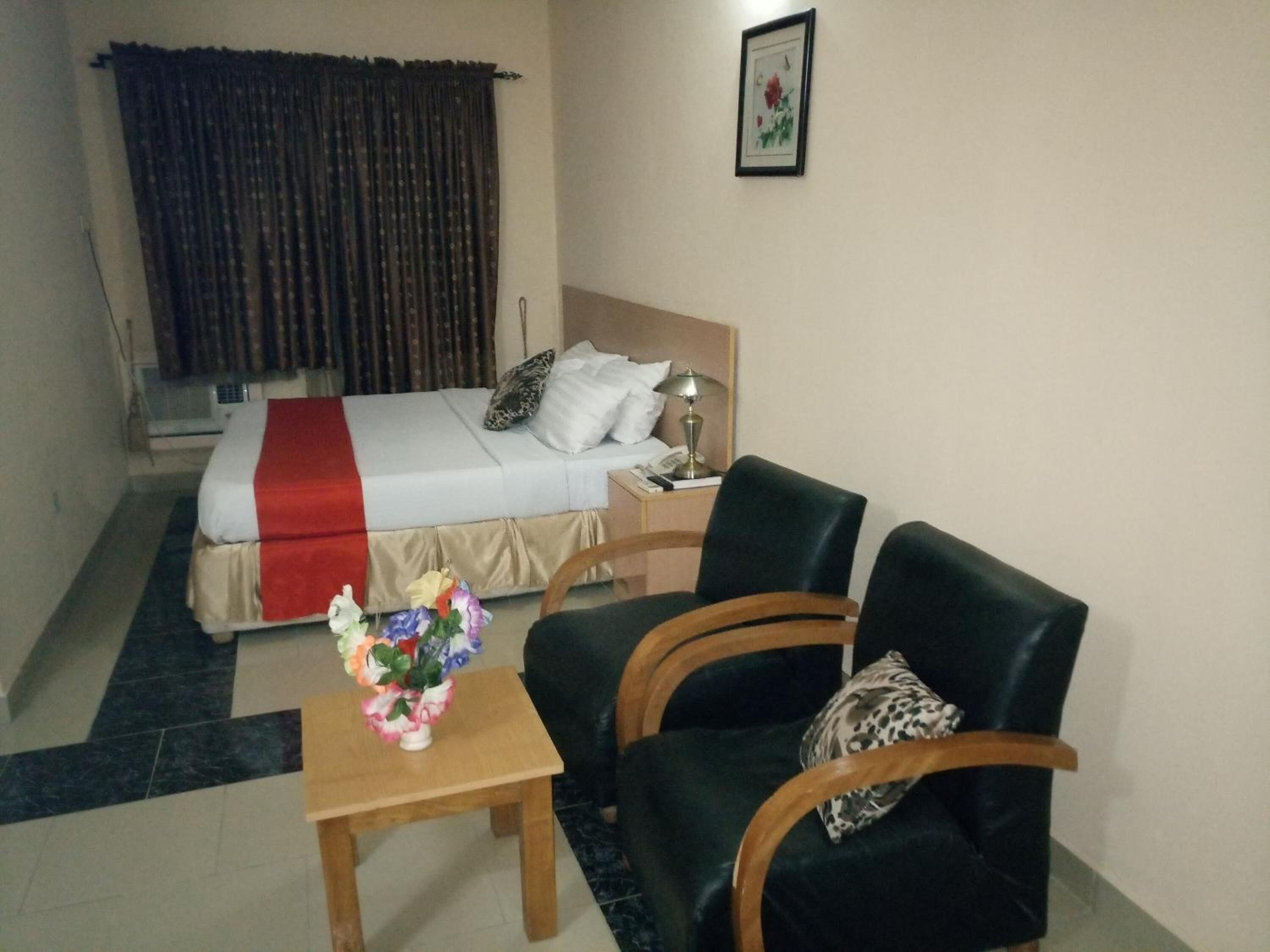 Une chambre d’hôtel avec un lit, deux chaises, une petite table sur laquelle est posé un vase de fleurs, une lampe et un tableau au mur.