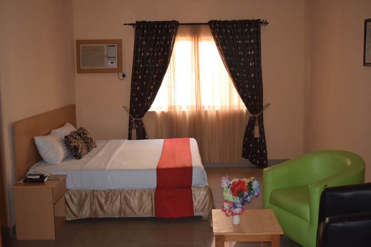 A hotel room with a bed, a nightstand, a green armchair, a small table, and a window with curtains.