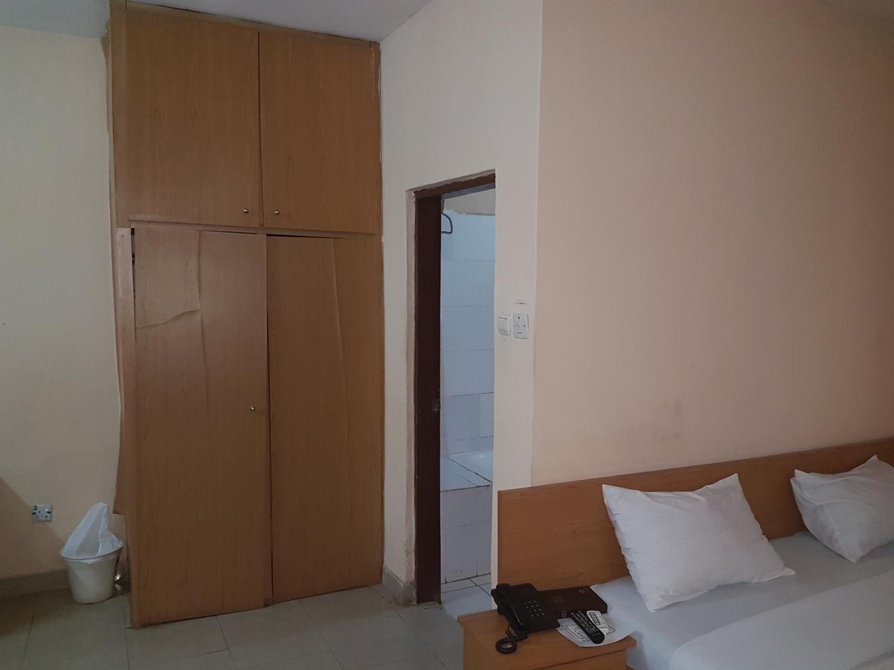 Quarto com guarda-roupa de madeira, cama com almofadas brancas, mesa de cabeceira com telefone e controle remoto, e lixeira.