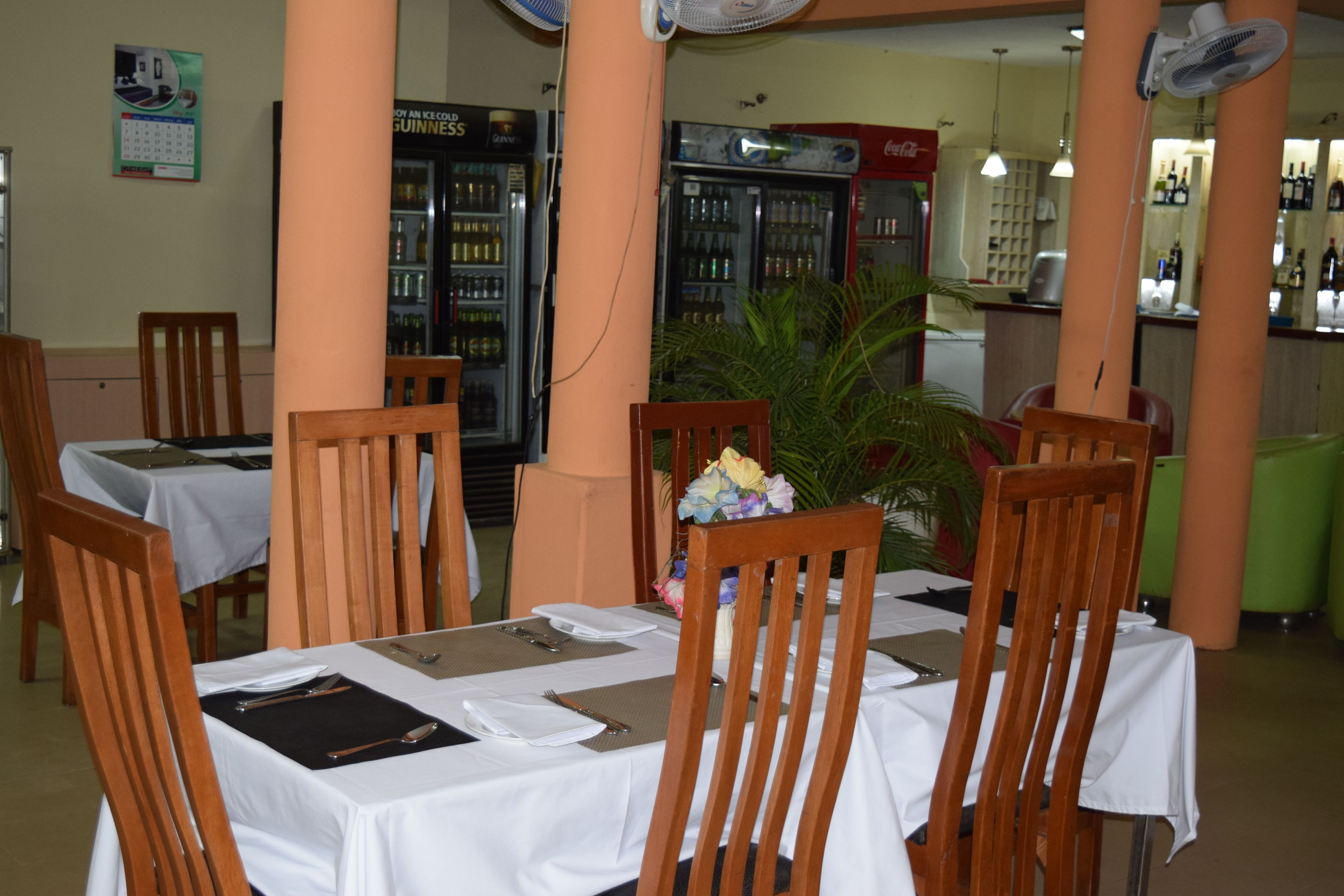 A restaurant with wooden chairs and tables, a plant, and refrigerators with beverages.