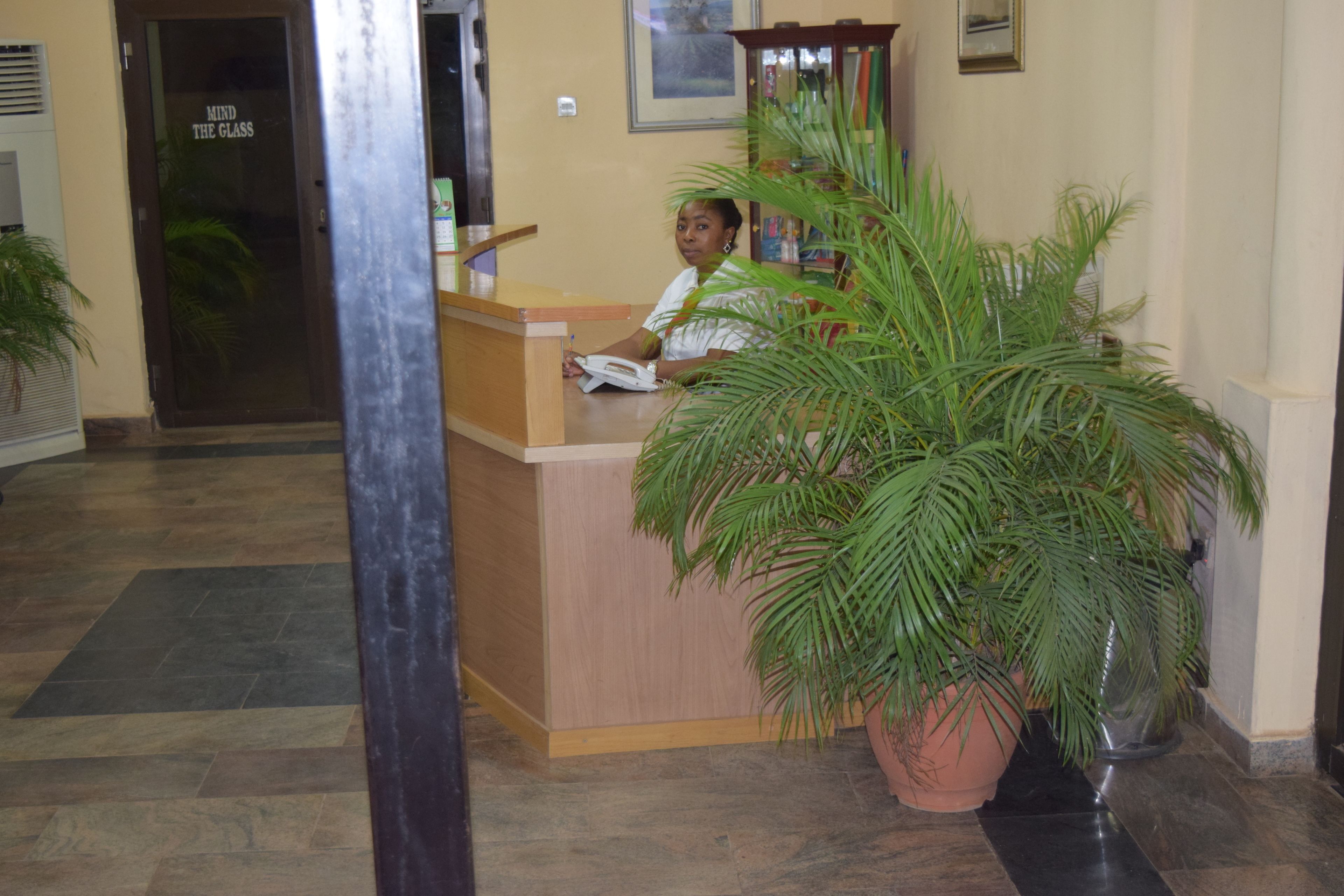 A reception desk with a receptionist, a large potted plant, and a door with a sign that reads "MIND THE GLASS".