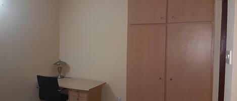 A small room with a wooden wardrobe, a desk with a chair, and a trash bin.