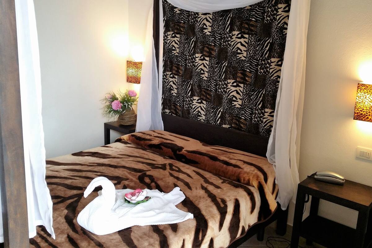 A bedroom with a zebra-patterned bedspread, a canopy bed, a bedside table with a lamp, and a wall-mounted light.