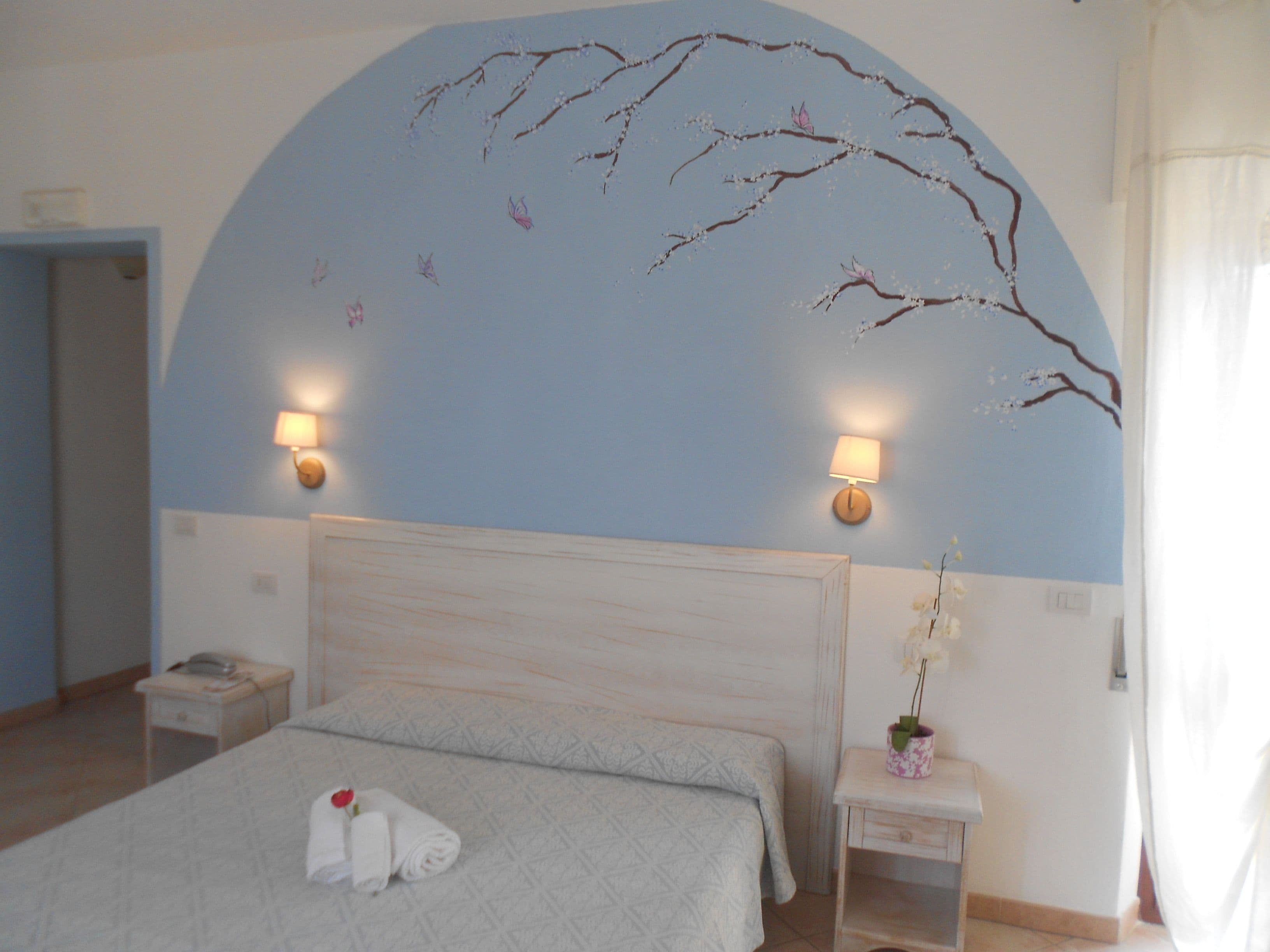 Un dormitorio con una cama grande, dos mesitas de noche, una mesita auxiliar, una lámpara de pared y una decoración mural.