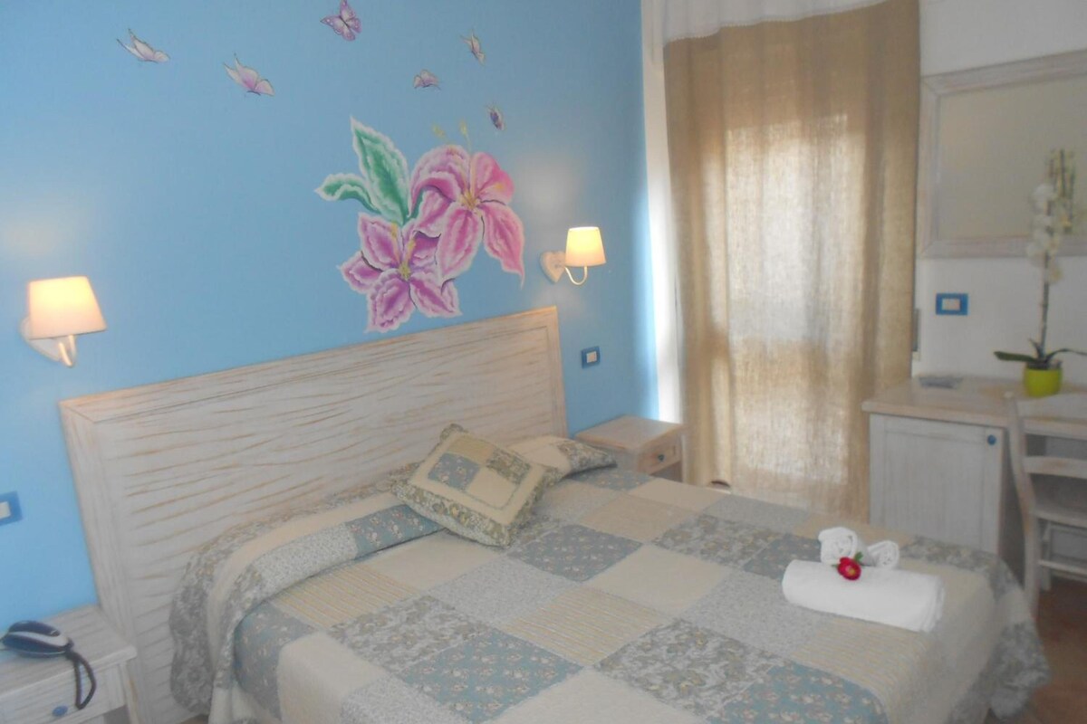 A bedroom with a bed, bedside lamps, a nightstand, a desk, a mirror, and a wall with a floral mural.