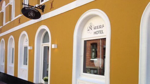 Un edificio amarillo con un letrero que dice "Mans Hotel". Se ven ventanas con marcos blancos y una puerta.