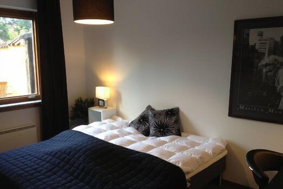 Un lit bien fait, avec une couette blanche et une couverture foncée, une lampe de chevet, un tableau encadré au mur et une fenêtre avec des rideaux.