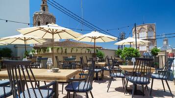 Un área de comedor al aire libre con mesas y sillas, sombrillas y vista a un edificio histórico al fondo.