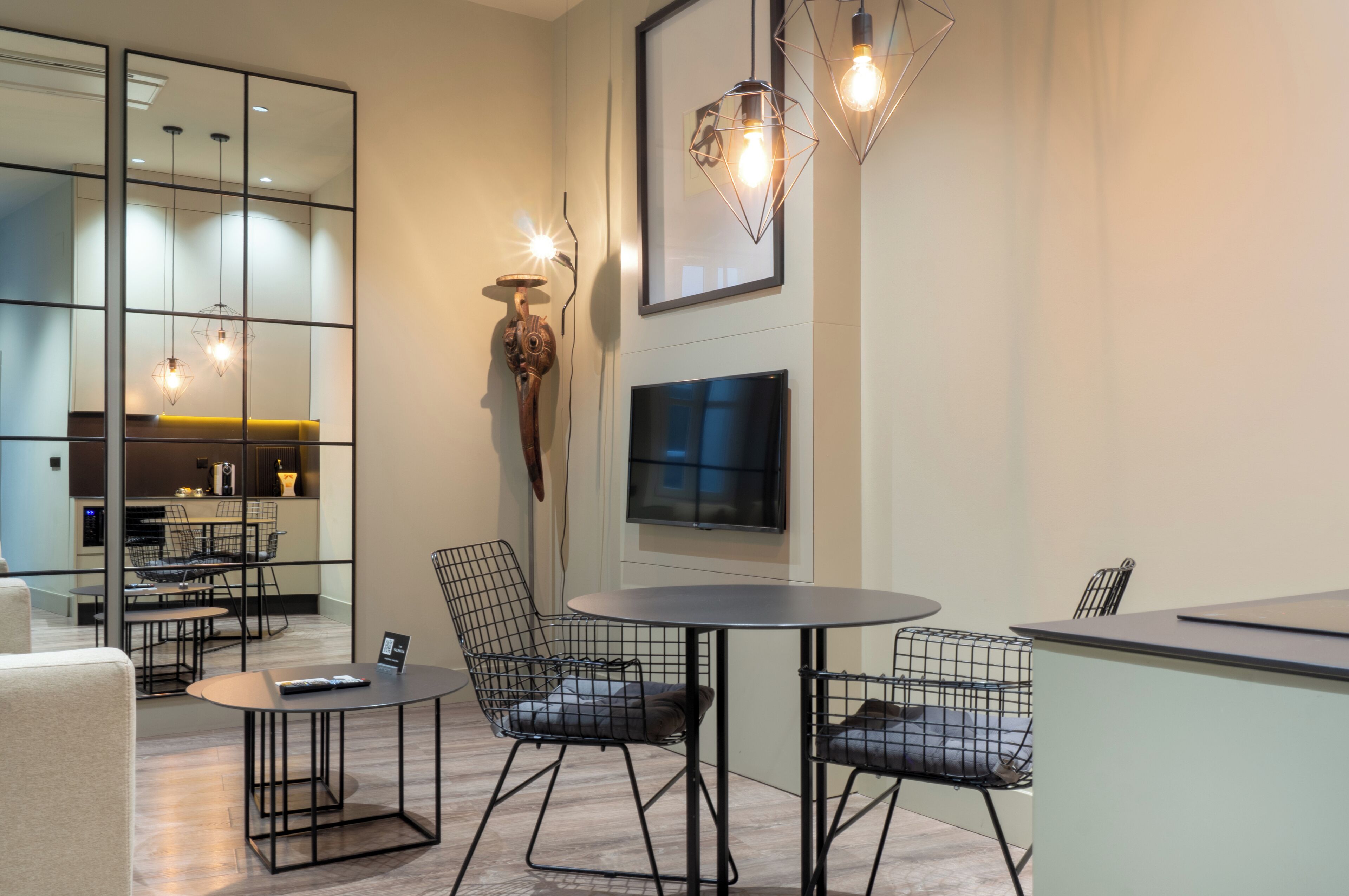 A modern interior with a round table, wire mesh chairs, a flat-screen TV, and pendant lighting.