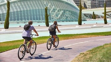 Two people riding bicycles on a paved path with a modern building and a body of water in the background.