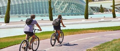 Two people riding bicycles on a paved path with a modern building and a body of water in the background.