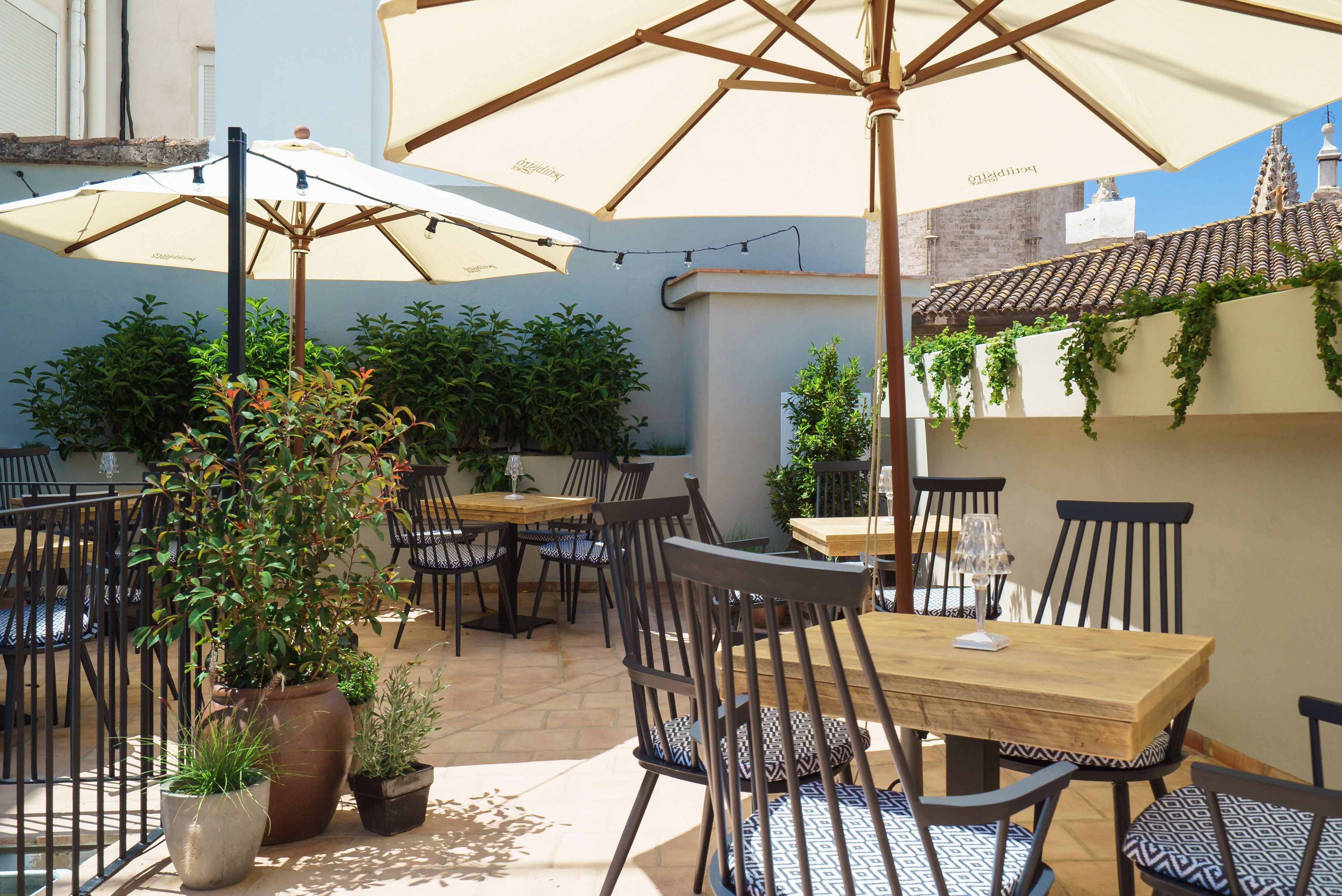 An outdoor patio with multiple tables and chairs, surrounded by potted plants and umbrellas.