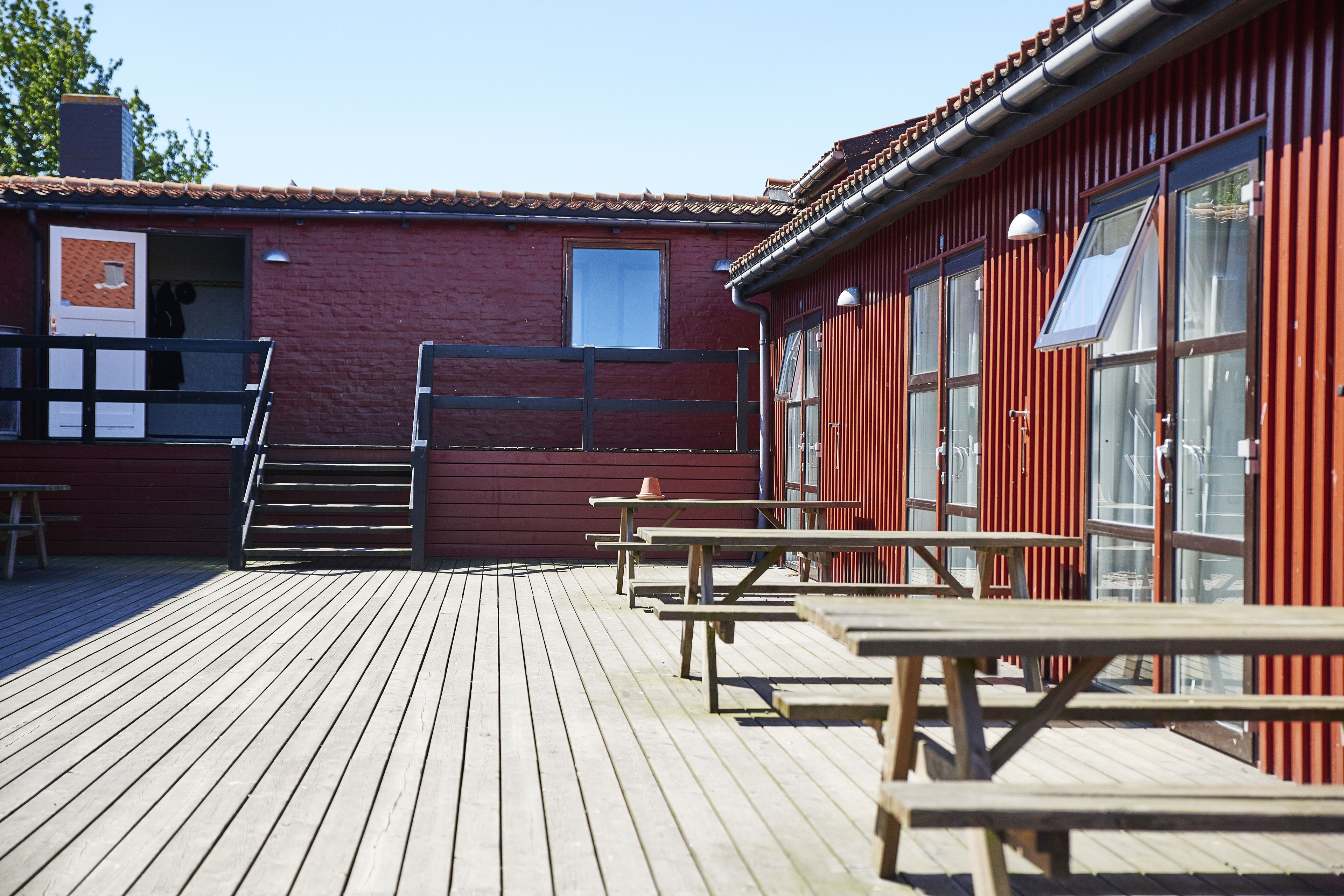 Una terraza de madera con mesas de picnic y un edificio rojo con ventanas grandes.