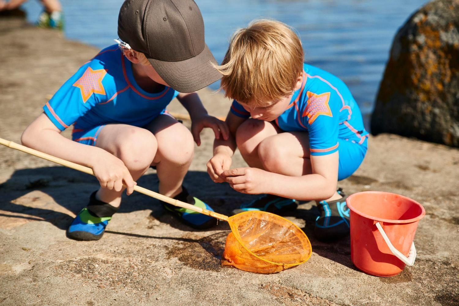 Two children playing near a body of water, one holding a net and a stick, and a red bucket nearby.