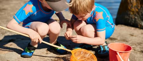Two children playing near a body of water, one holding a net and a stick, and a red bucket nearby.