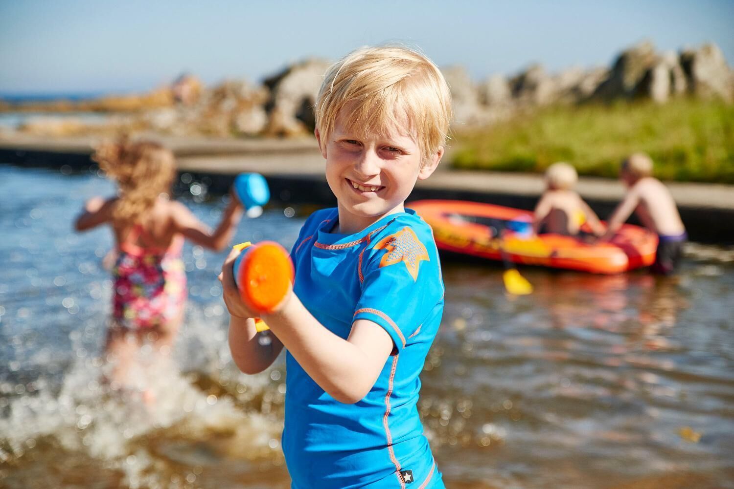 A child in a blue shirt holding a toy in a body of water with other children and inflatable rafts in the background.