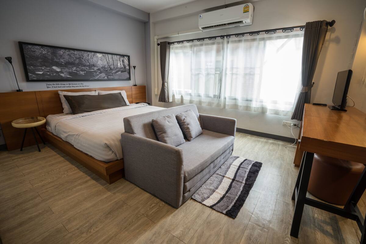 A bedroom with a bed, a sofa, a desk, a chair, a rug, a picture on the wall, and a window with curtains.