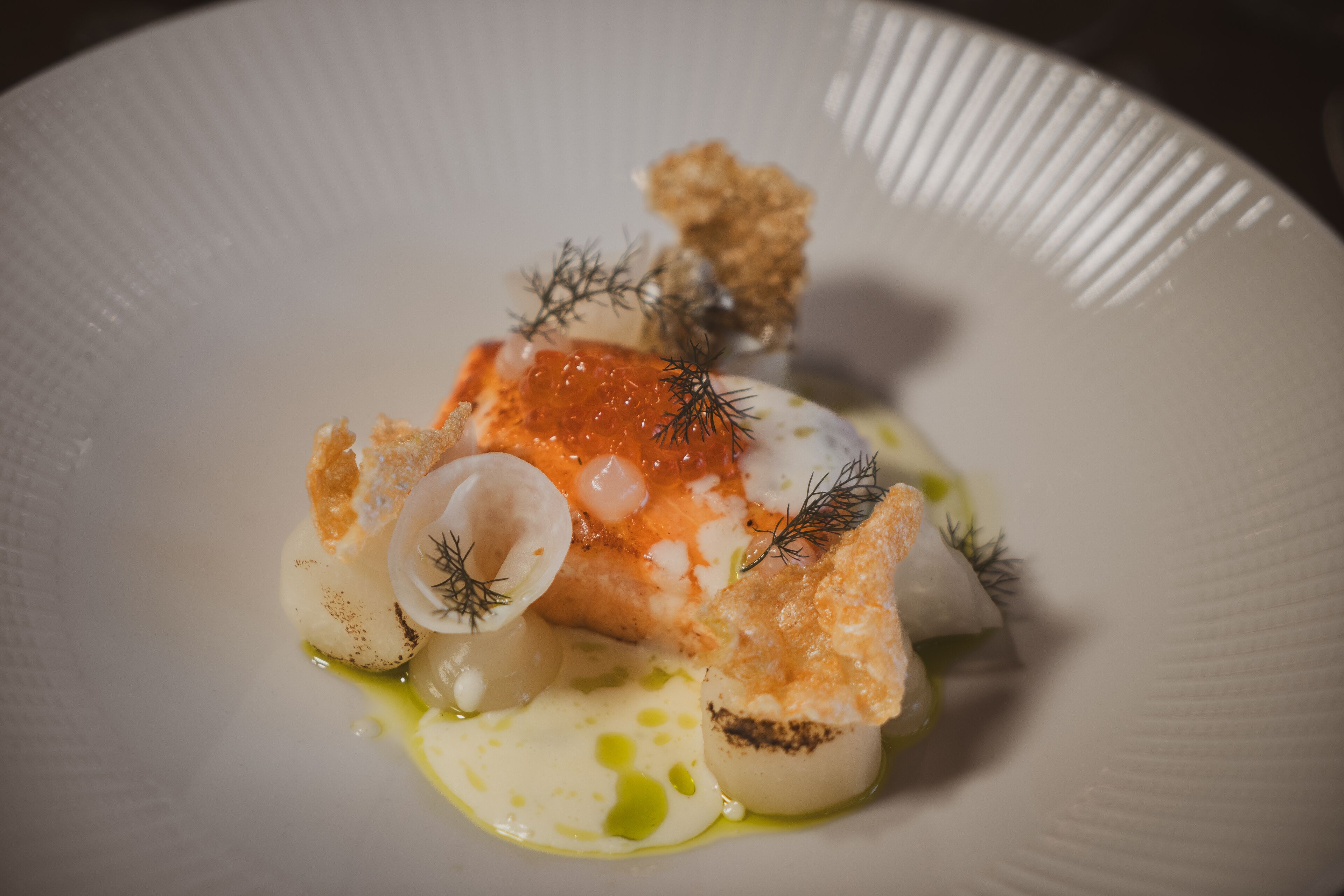 A white plate with a piece of fish topped with orange caviar, garnished with dill and served with a sauce.