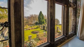 Vista a través de una ventana con marco de piedra, que muestra un jardín bien cuidado con césped circular, senderos de piedra y diversos árboles.