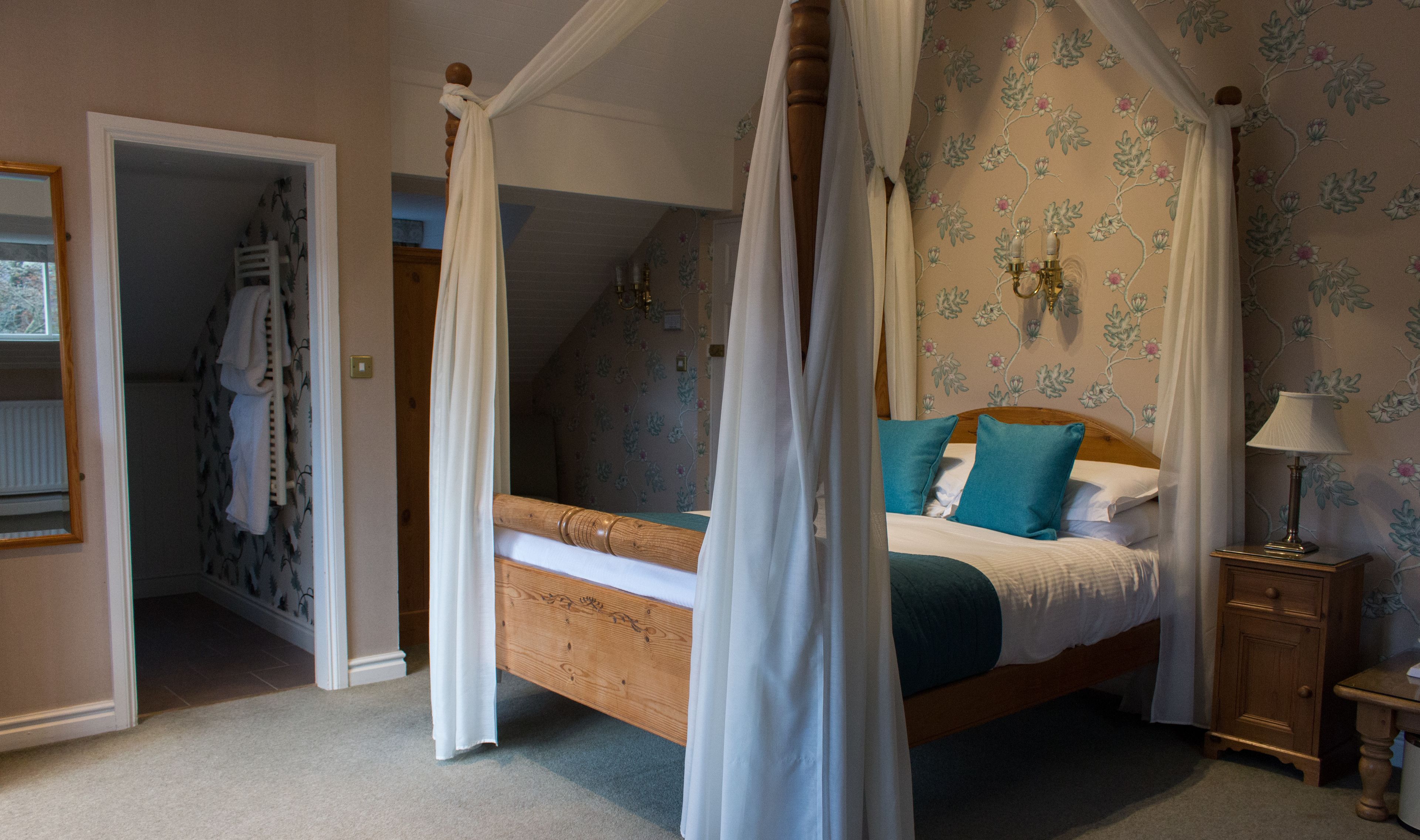 A four-poster canopy bed with a floral wallpaper, a wooden nightstand, and a night lamp.