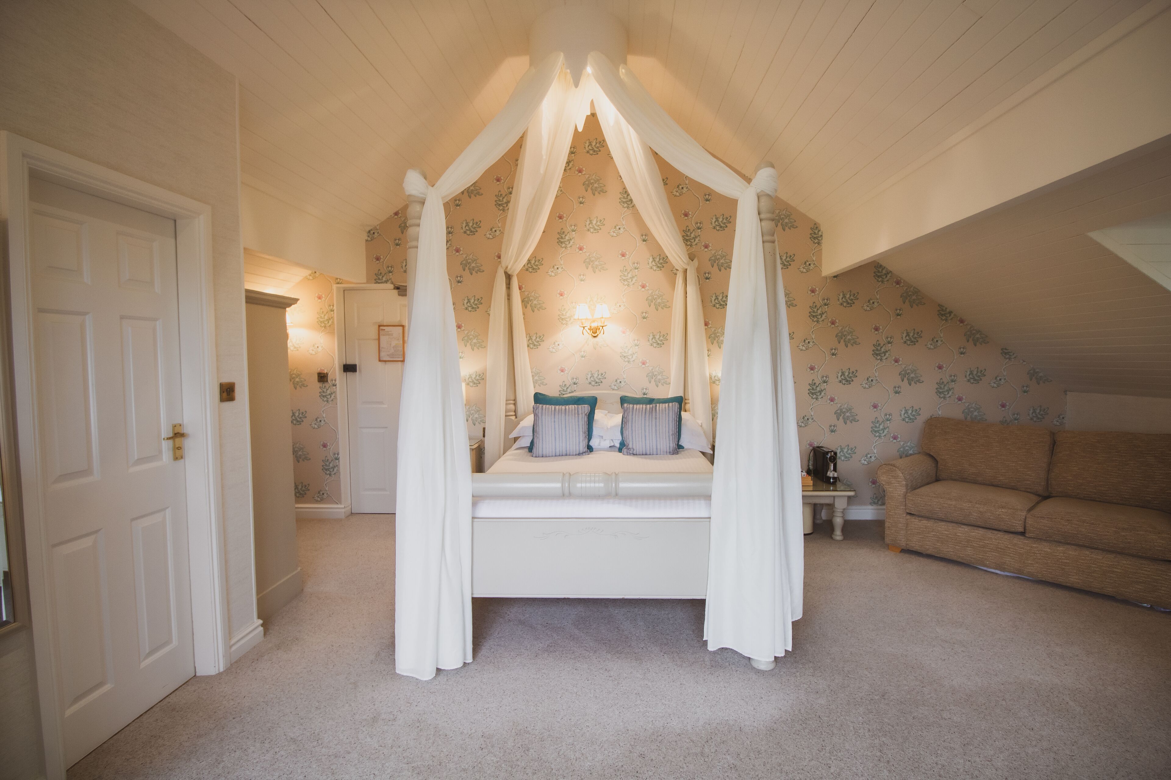 A bedroom with a canopy bed, a white door, a beige sofa, and floral wallpaper.