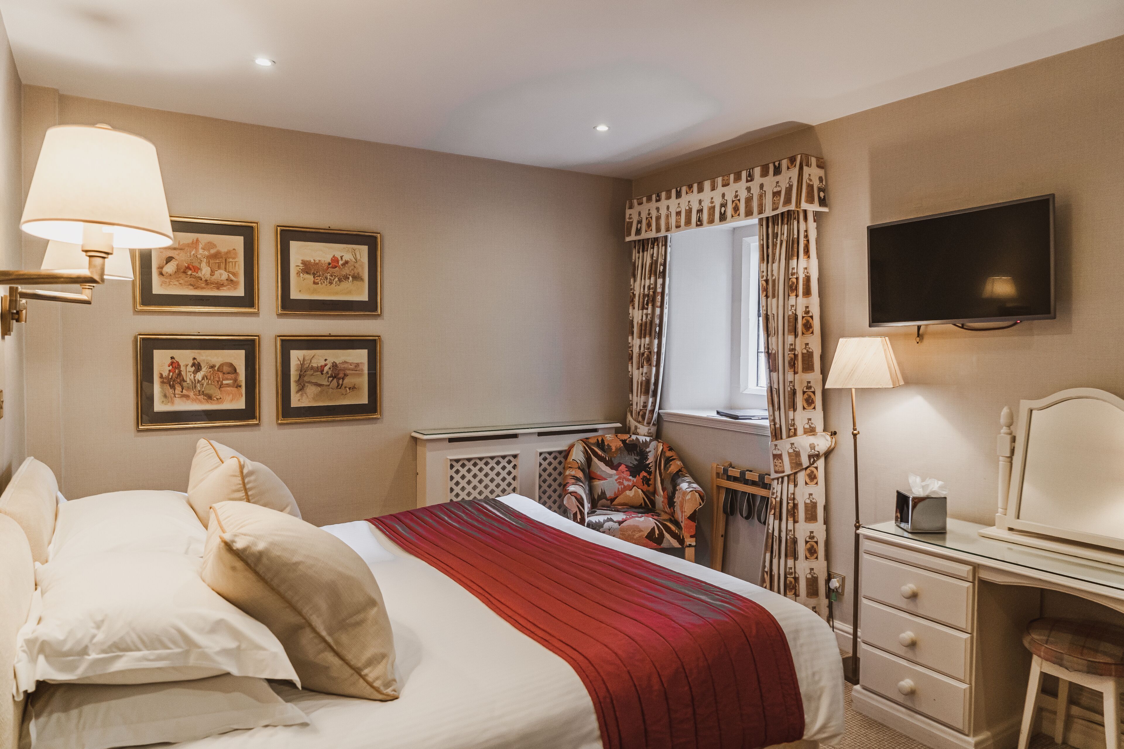 A hotel bedroom with a bed, a desk, a chair, a TV, and framed pictures on the wall.