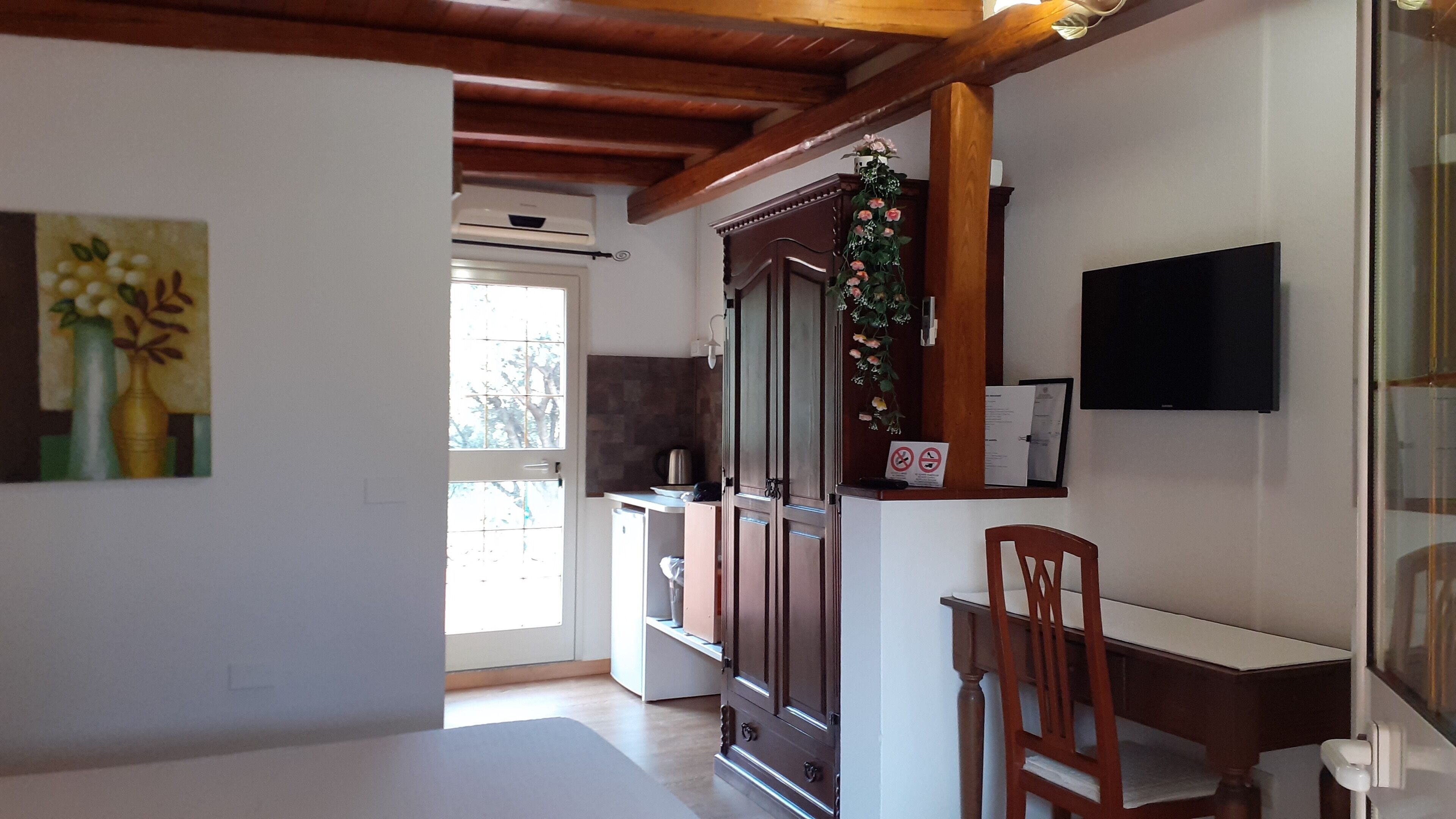 A room with a wooden ceiling, a TV mounted on the wall, a desk with a chair, and a cabinet with a decorative plant.