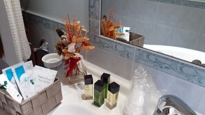 A bathroom with a white sink, a mirror, a vase with dried flowers, a basket with toiletries, and a plastic bag.