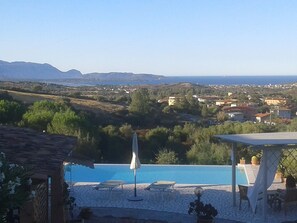 A pool area with lounge chairs and an umbrella, overlooking a coastal town with houses and mountains in the distance.