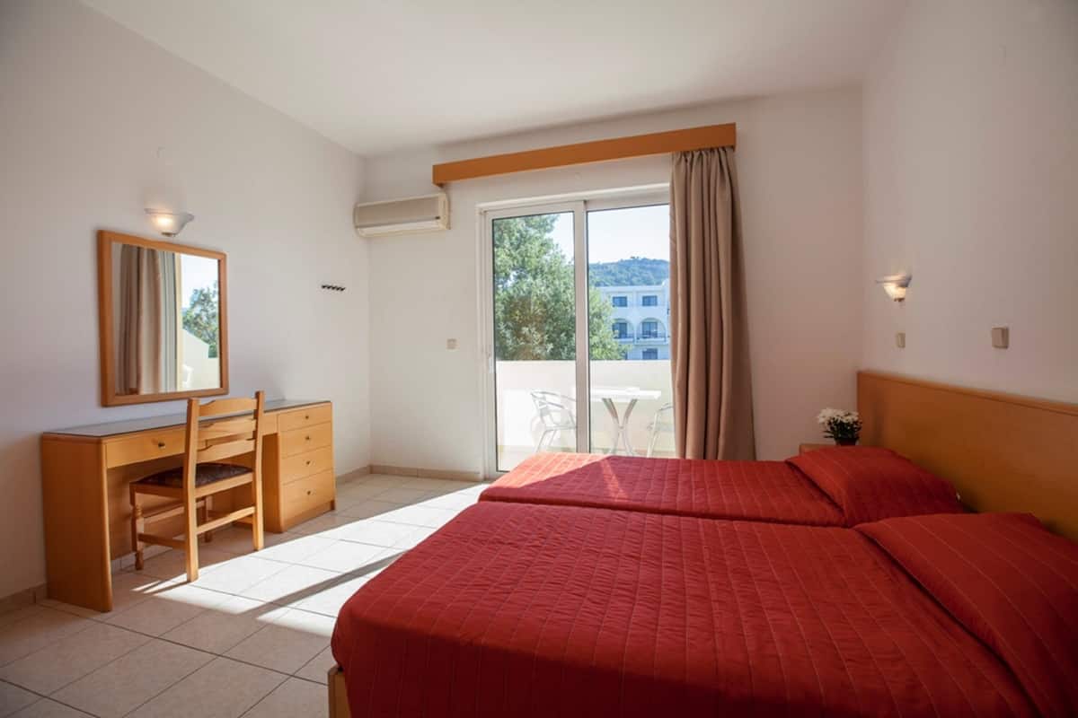 A bedroom with two beds, a desk, a chair, a mirror, and a sliding glass door to a balcony.