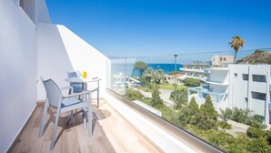 A balcony with a clear view of a pool and buildings, a table with chairs, and a glass railing. - Moscha Hotel (Rhodes)