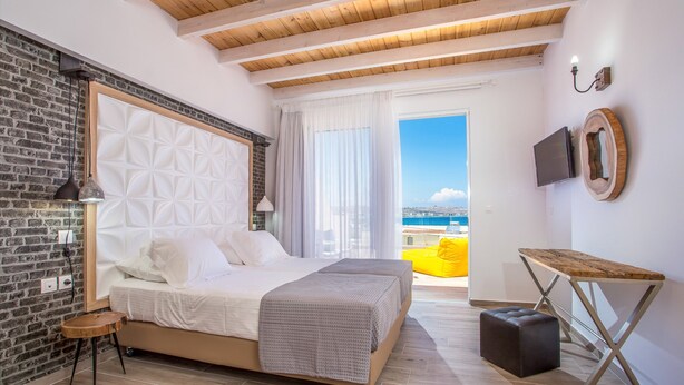 A modern bedroom with a large bed, a small side table, a wooden bench, and a TV on the wall. - Moscha Hotel (Rhodes)