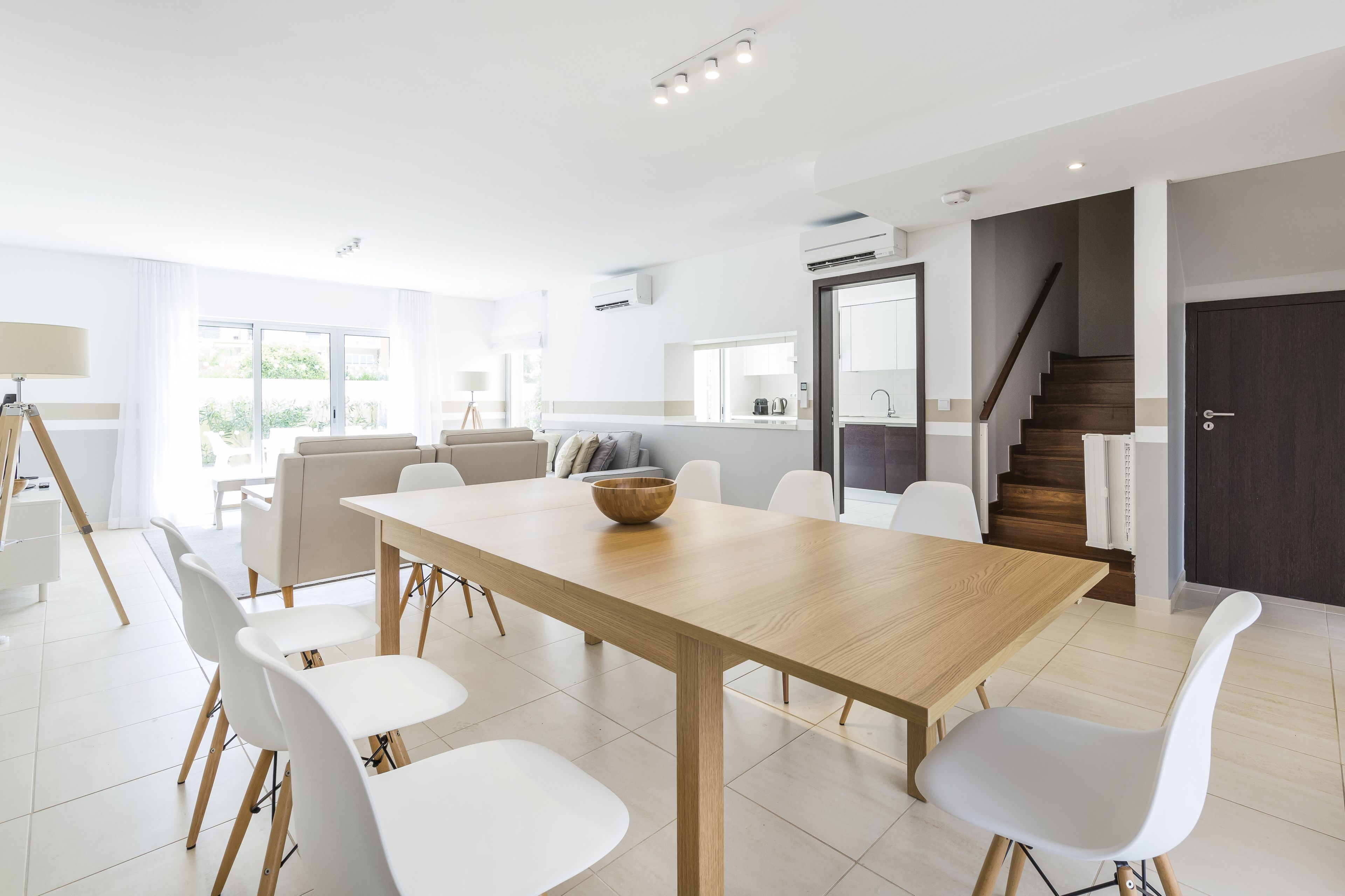 A modern dining area with a wooden table and white chairs, a kitchen in the background, and a staircase leading to an upper level.