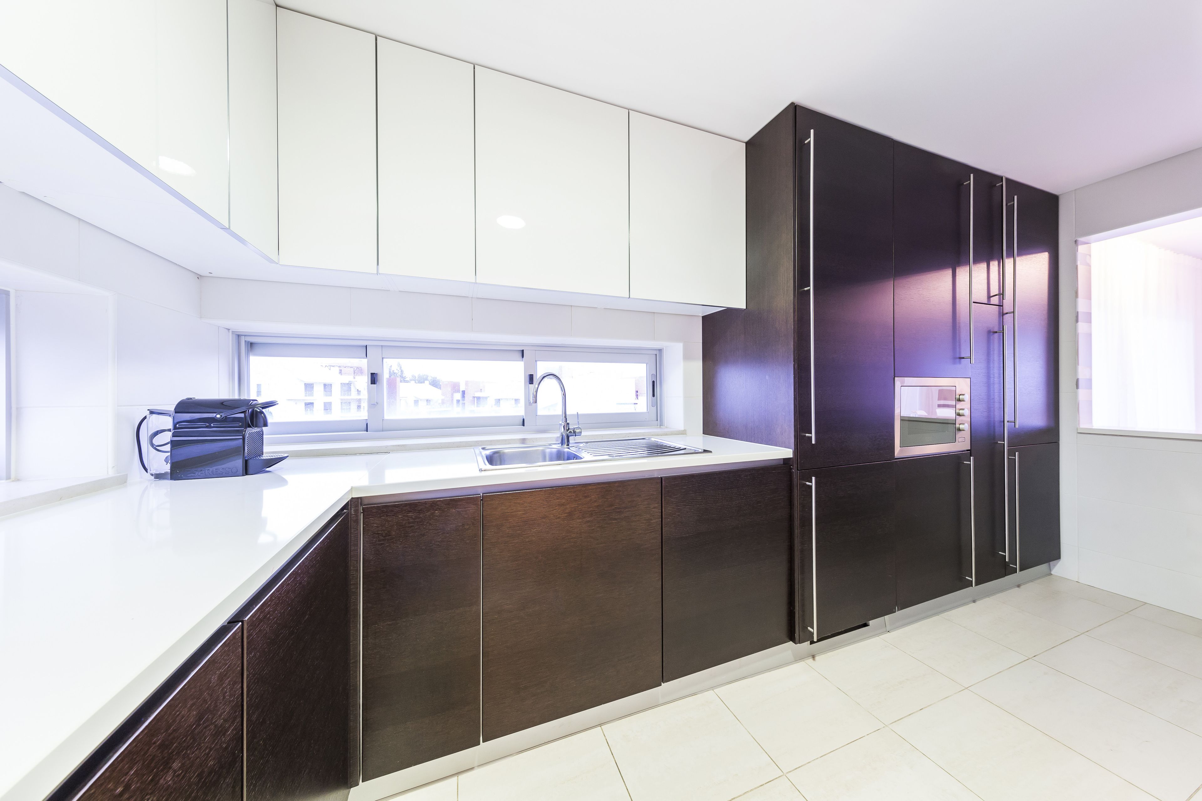 A modern kitchen with a white countertop, dark cabinets, and a window providing natural light.