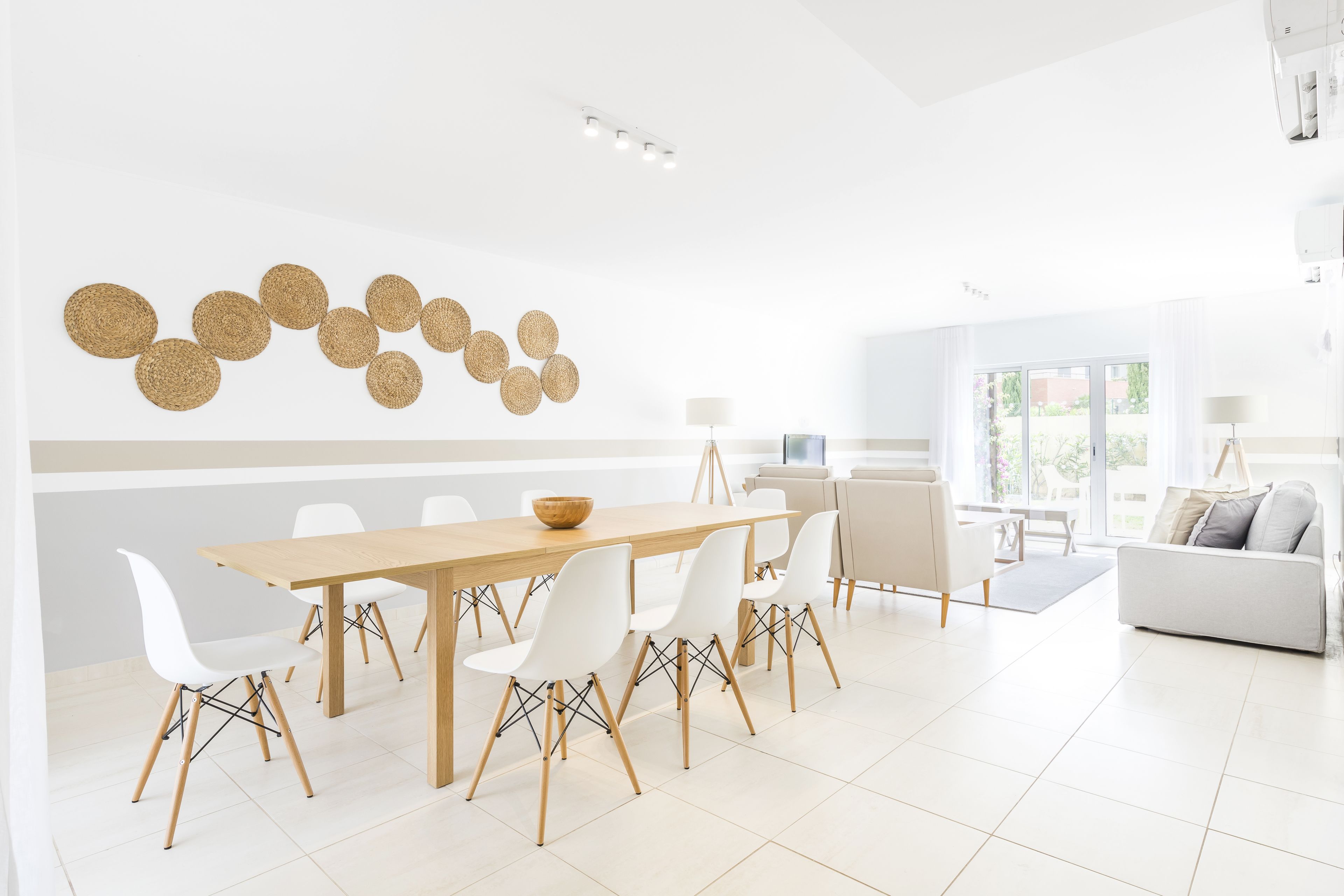 A modern dining area with a wooden table and white chairs, a decorative wall hanging, and a view of the outdoors through large windows.
