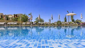 En swimmingpool med kakelbeklädda kanter, solstolar och parasoller, omgiven av byggnader och träd.