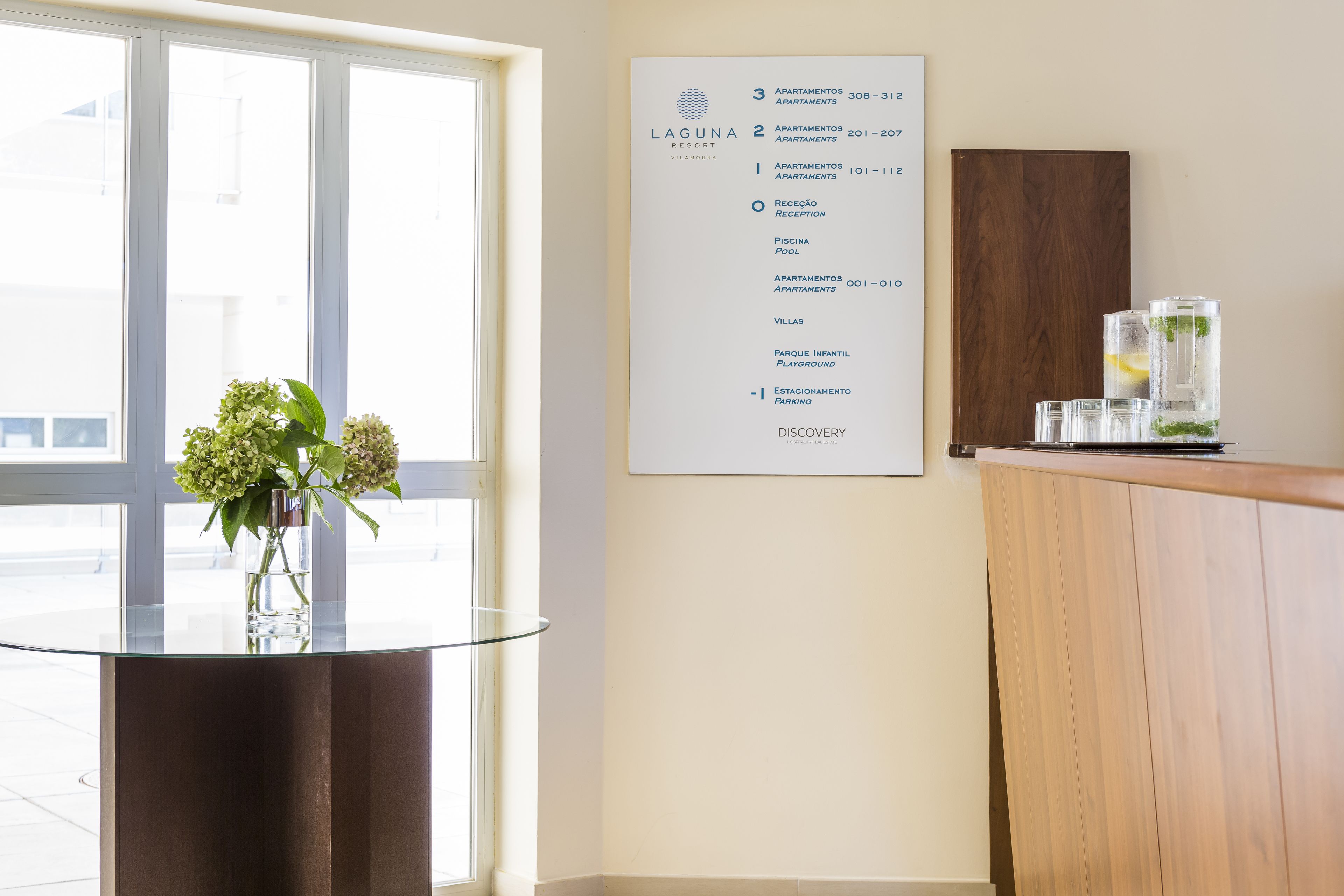 Zona de recepción de un hotel con una mesa de vidrio, un jarrón con flores y un cartel que enumera servicios como apartamentos, piscina y aparcamiento.