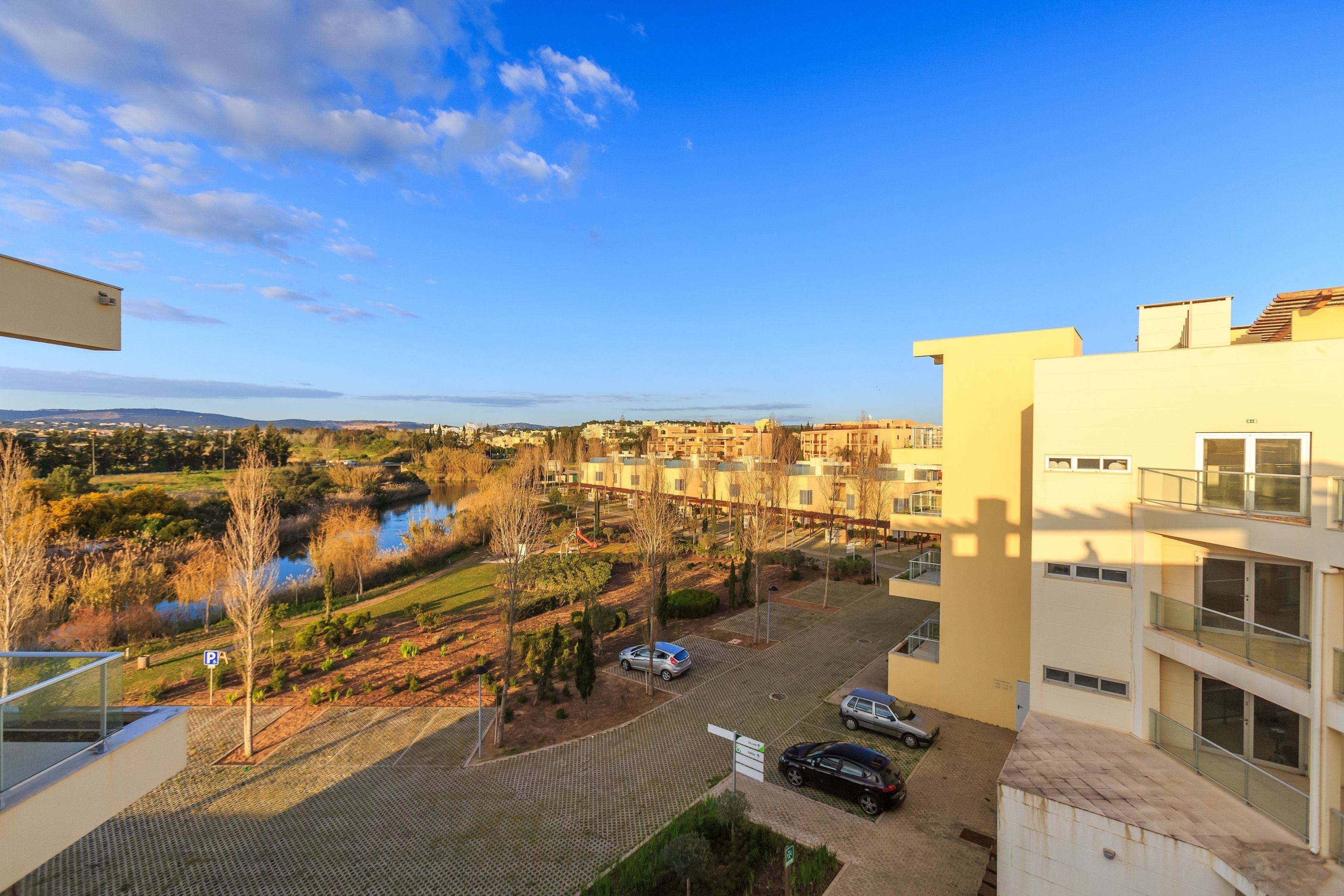 A residential area with a paved courtyard, parked cars, and a view of a lake and distant buildings.