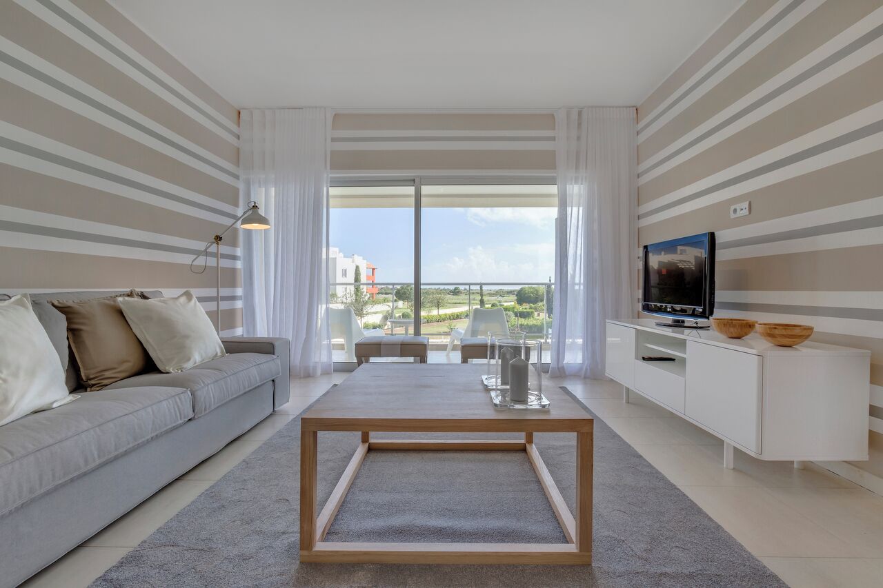 A modern living room with a grey sofa, a wooden coffee table, a TV on a white cabinet, and a balcony with a view of buildings and greenery.