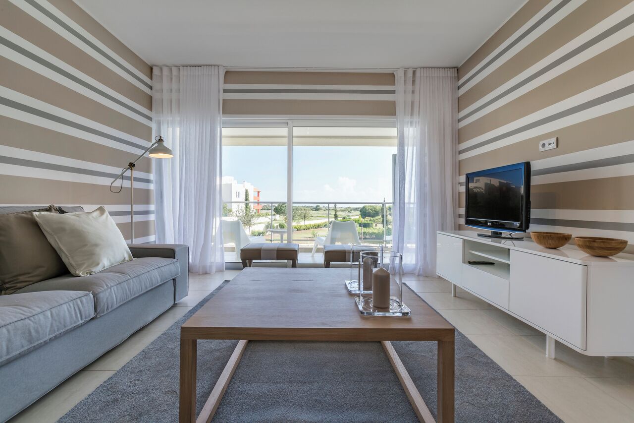 A modern living room with a sofa, a coffee table, a TV, and a balcony with a view of the city.