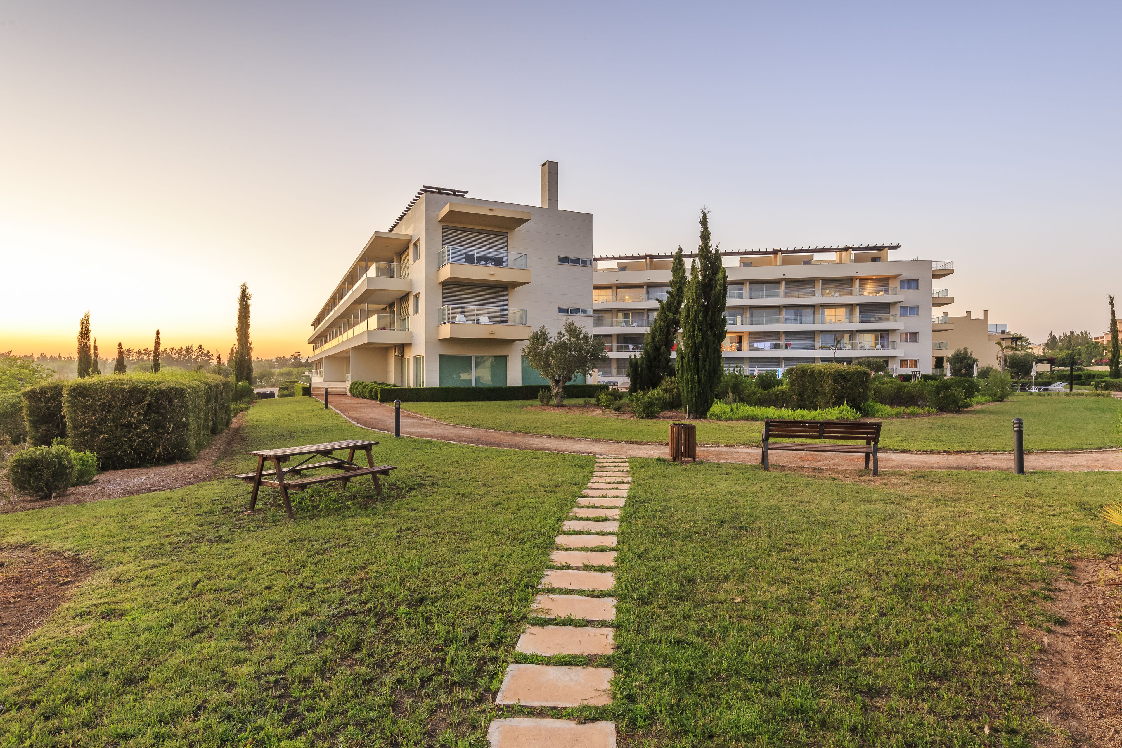 Un edificio moderno con varios balcones, un camino pavimentado y una zona ajardinada con un banco.