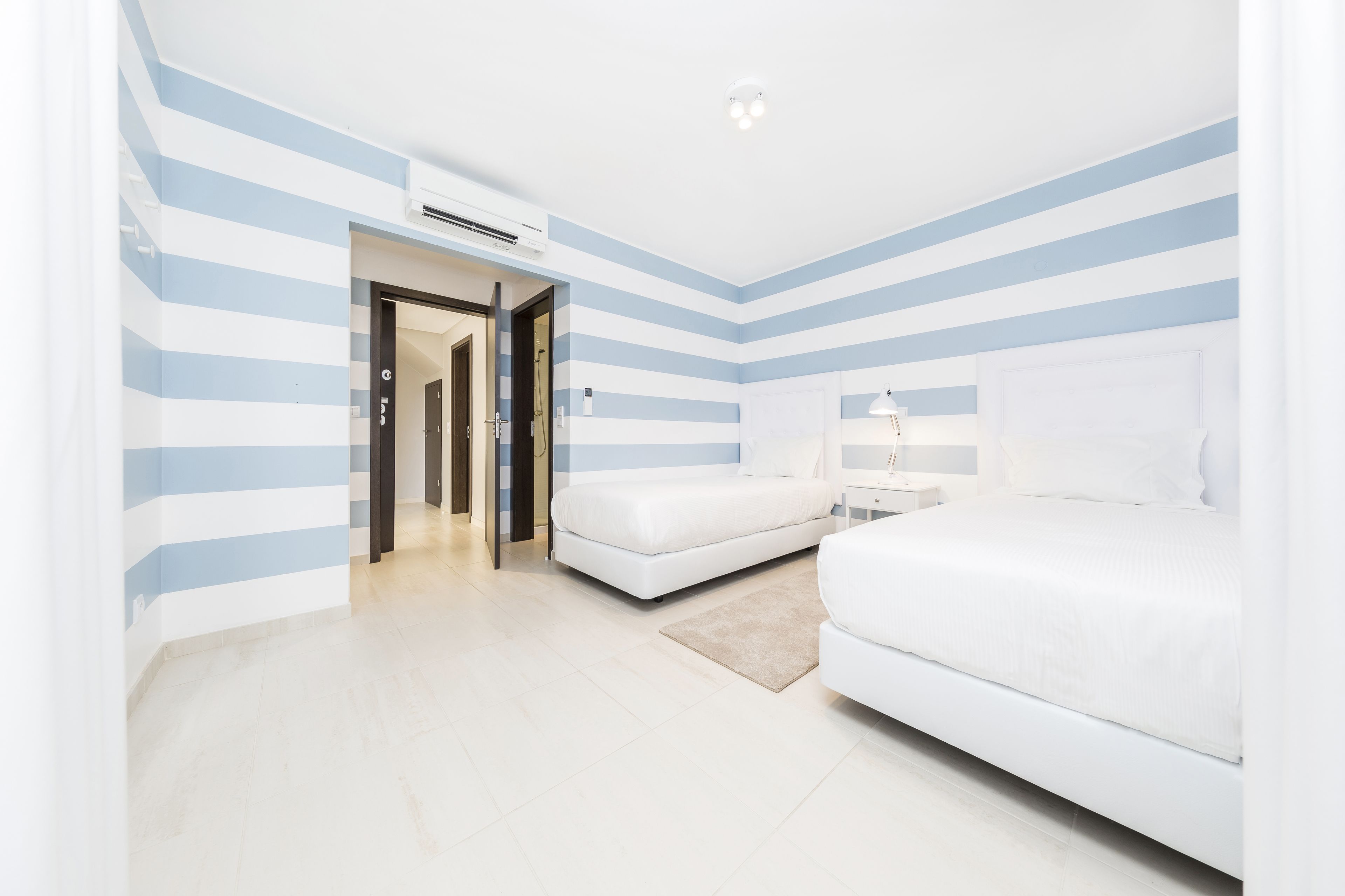 A hotel room with a striped wall design, a bed, a bedside table with a lamp, and a door leading to another room.