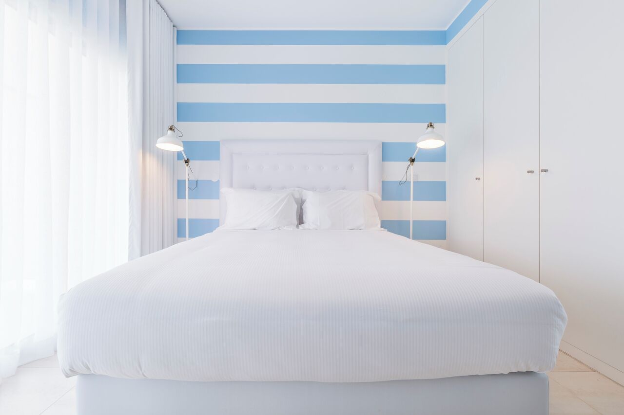 A large bed with white bedding and a blue-striped wall behind it. There are two bedside lamps on either side of the bed.