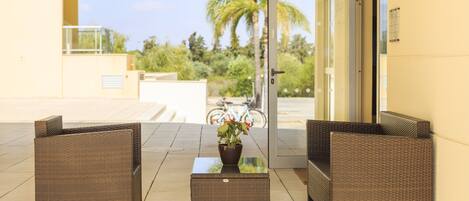 A reception area with wicker furniture, a glass-top table with a potted plant, and a view of palm trees and a bicycle in the background.