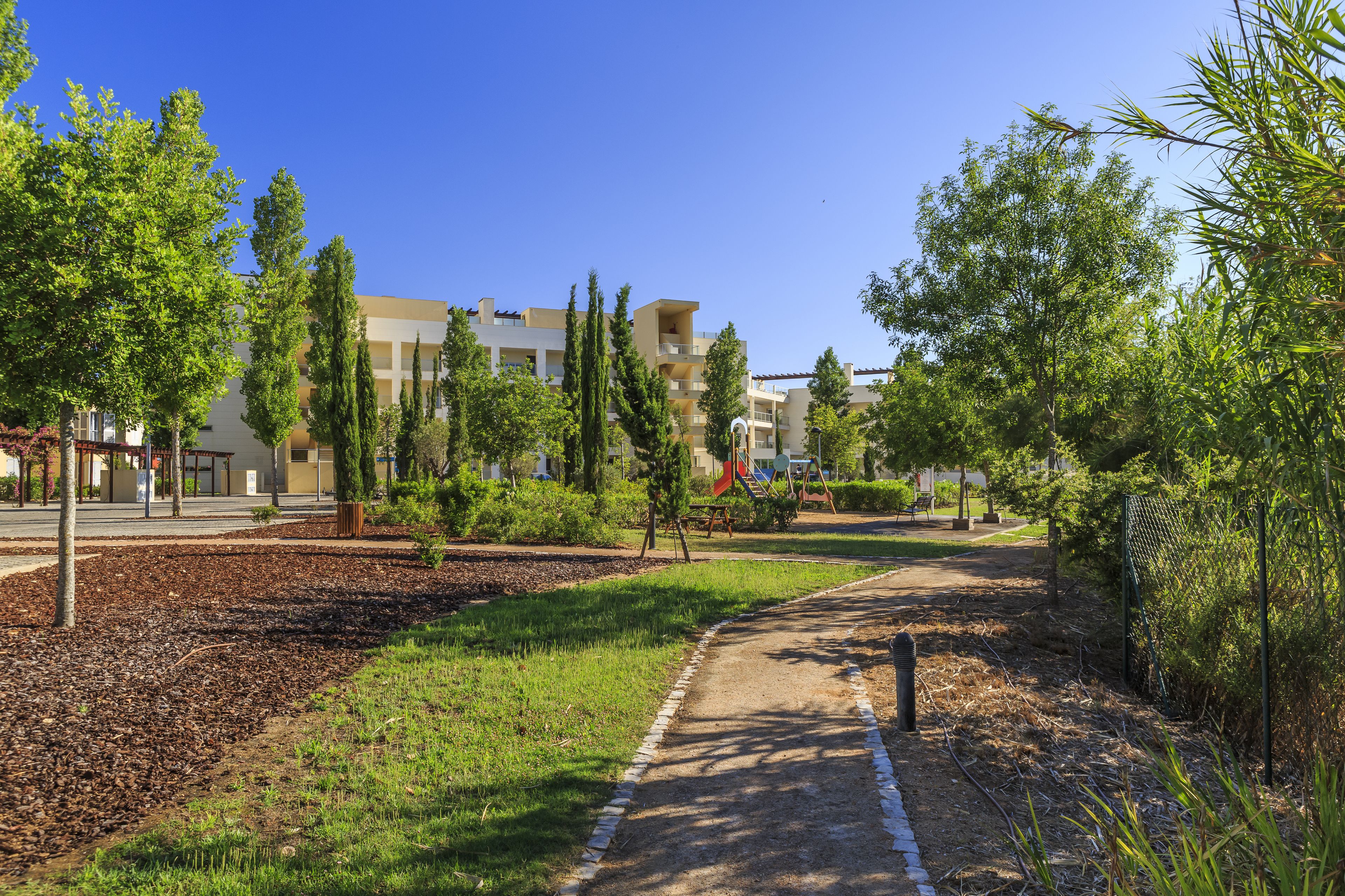 A pathway leading to a playground with a slide, surrounded by trees and a building with multiple balconies.
