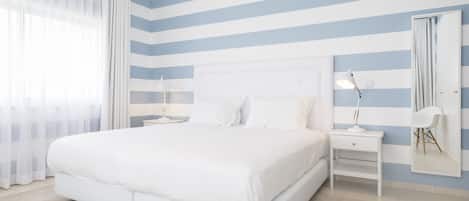 Une chambre à coucher avec un grand lit, deux tables de chevet blanches, une table de nuit, un miroir et une fenêtre ornée de rideaux légers.